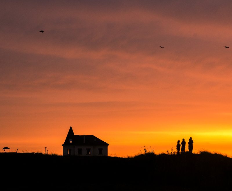 A house with a pointed tower and three people silhouetted on a ridge against an orange sunset, birds overhead.