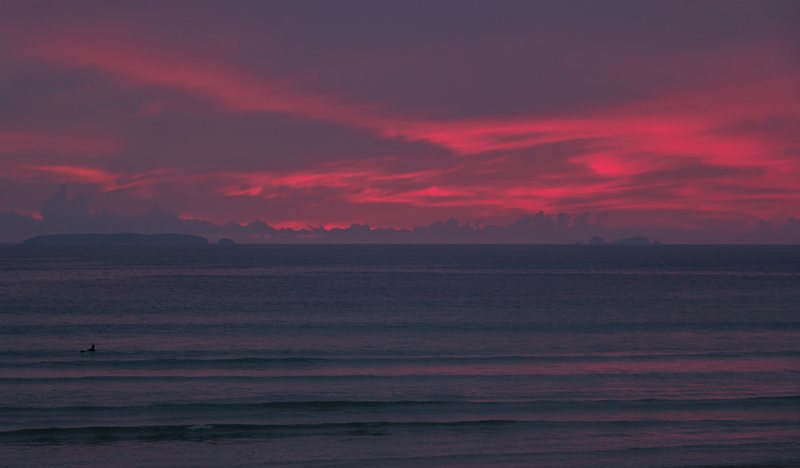 Streaks of red cloud over a calm purple sea at dusk, a lone surfer in the water and a low island on the horizon.