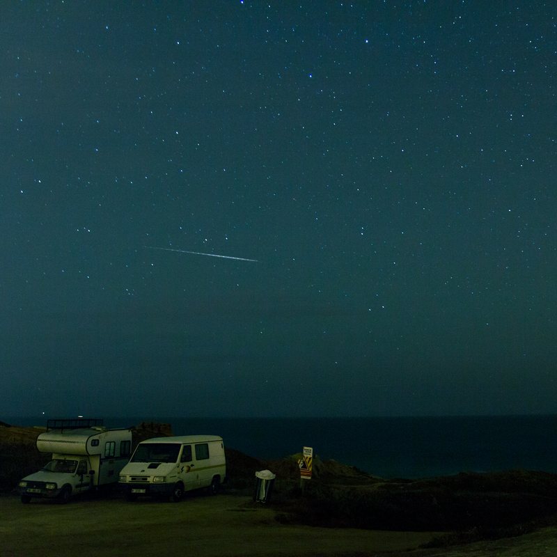 A shooting star crosses a teal night sky full of stars above two camper vans parked by the sea.