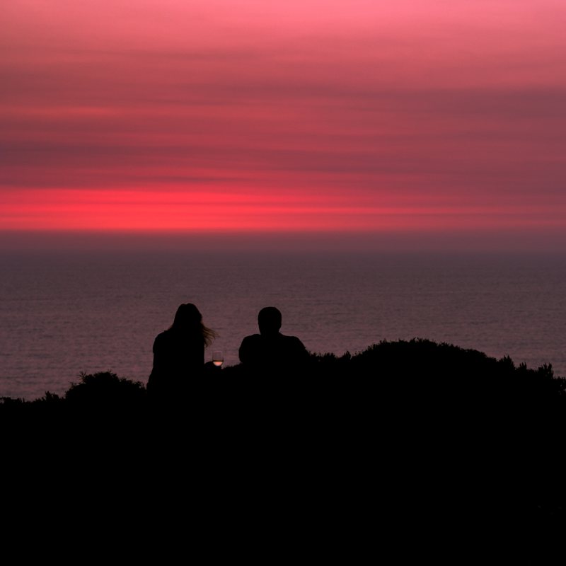 Two people sit silhouetted on a dark hilltop above the sea, under bright bands of red and pink sky.