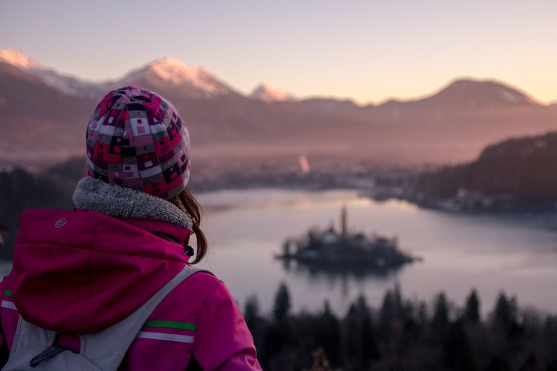 A child in a pink jacket and knitted hat looks down at a lake with a small island church, pink morning light on the mountains.