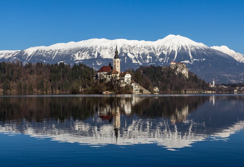 An island church and its reflection in a calm blue lake, snow-covered mountains behind.