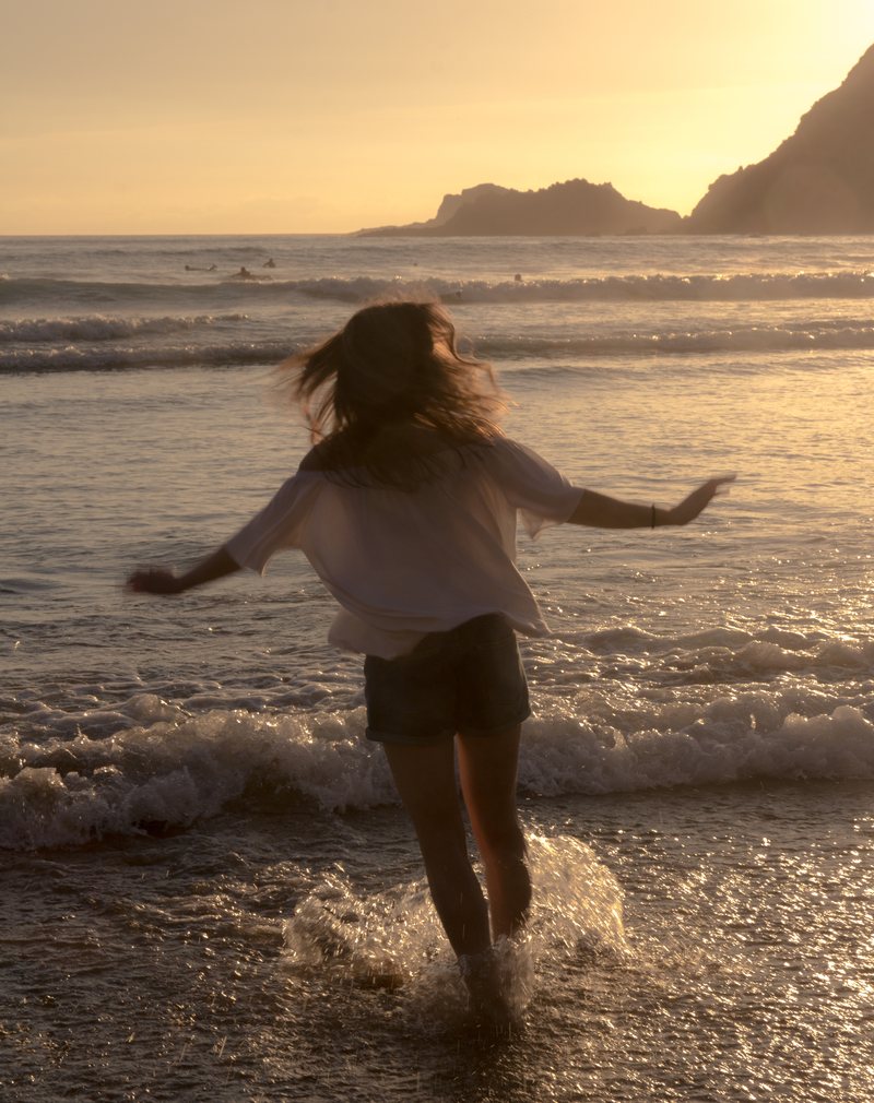 A woman with windswept hair runs into the surf at golden hour, arms out, water splashing around her legs.