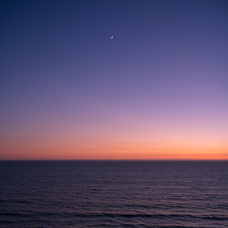 A thin crescent moon above a calm sea, the sky fading from deep blue to violet to an orange horizon.