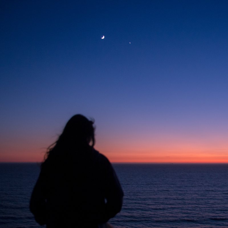 A silhouetted woman looks out to sea under a crescent moon, the horizon glowing orange beneath a deep blue sky.
