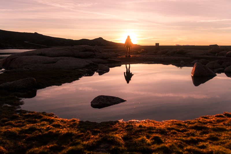 A figure stands on rocks against the setting sun, reflected in a still tide pool surrounded by glowing seaweed.
